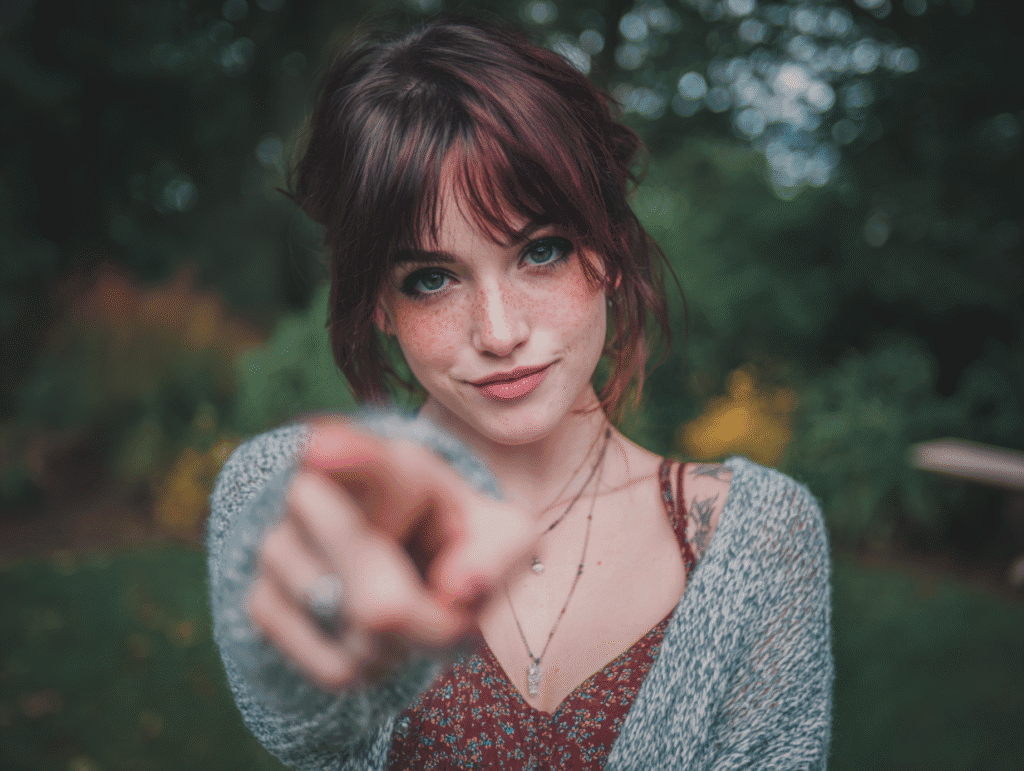 Smiling young woman with red hair and freckles pointing toward the camera in an inviting gesture outdoors